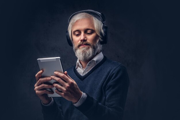 Studioportrait eines hübschen älteren Mannes, der ein Tablet mit Kopfhörern über einem dunklen Hintergrund verwendet.