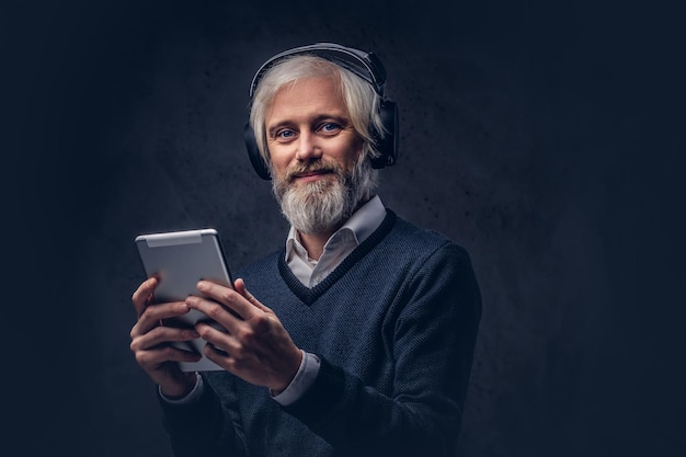 Studioportrait eines hübschen älteren Mannes, der ein Tablet mit Kopfhörern über einem dunklen Hintergrund verwendet.