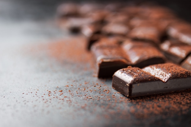 Stücke Schokolade Tablette auf einem schwarzen Holztisch und Kakao bestreut auf Top