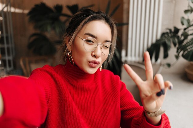 Stylische Asiatin mit Brille, goldenen Ohrringen und rotem Outfit zwinkert, macht Selfie und zeigt Peace-Zeichen