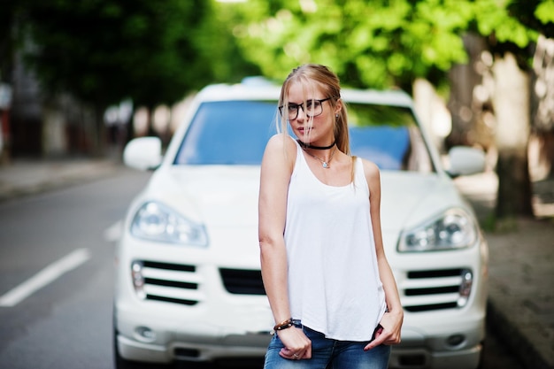 Stylische blonde Frau mit Jeansbrille und weißem Hemd gegen Luxusauto Urbanes Modellporträt der Mode