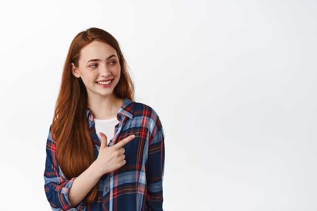 Stylische junge Frau mit langen roten Haaren, zeigt und schaut direkt auf leeren Raum, Werbetext, lächelt und lacht entspannt, weißer Hintergrund