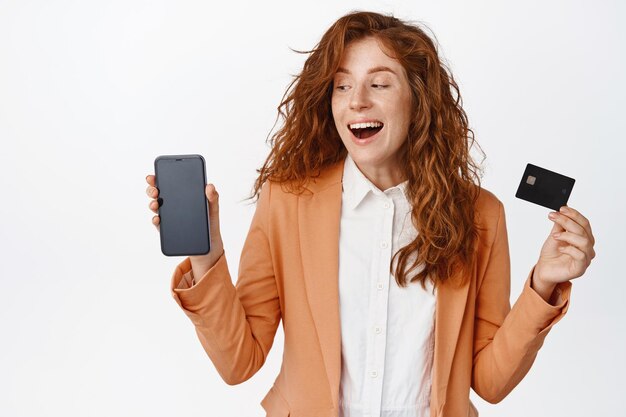 Stylische junge Geschäftsfrau mit rotem lockigem Haar, die Handybildschirm und Kreditkarte zeigt, lacht und lächelt zufrieden mit weißem Hintergrund