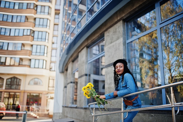 Stylische, modische afroamerikanische Frauen in Jeans und schwarzer Baskenmütze mit gelbem Blumenstrauß posierten im Freien an sonnigen Tagen vor blauem, modernem Gebäude