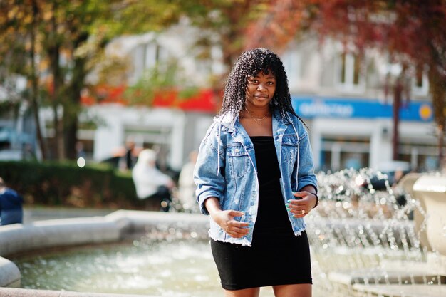 Stylische, trendige, lockige Afro-Frankreich-Frau posierte am Herbsttag in Jeansjacke Schwarzafrikanisches weibliches Modell gegen Brunnen