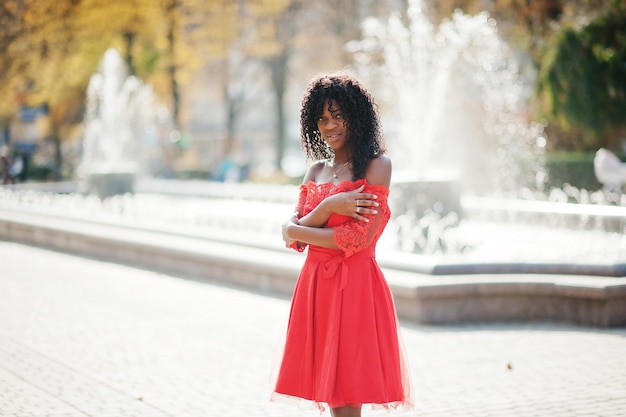 Stylische, trendige, lockige Afro-Frankreich-Frau posierte am Herbsttag in rotem Kleid Schwarzafrikanisches weibliches Modell gegen Brunnen