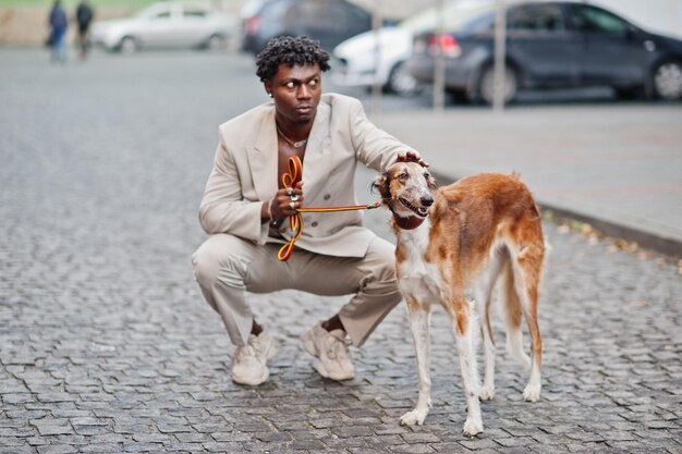 Stylischer Afro-Mann in beigem Oldschool-Anzug mit russischem Borzoi-Hund Modischer junger afrikanischer Mann in Freizeitjacke auf nacktem Oberkörper