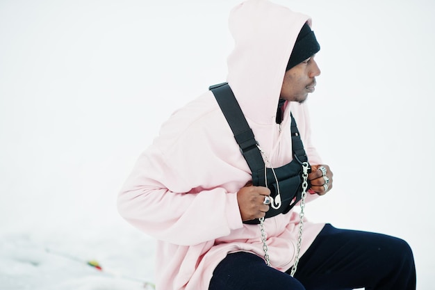 Stylischer afroamerikanischer Mann im urbanen Stil in rosafarbenem Hoodie, der im Winter am zugefrorenen See posiert