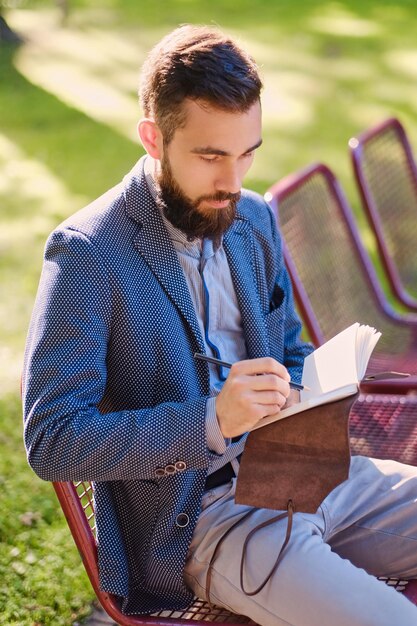 Stylischer bärtiger Mann, der eine Nachricht an einen Notizblock in einem Park schreibt.