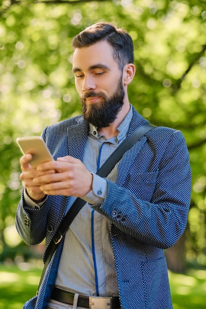 Stylischer bärtiger Mann in blauer Jacke mit Smartphone in einem Park.