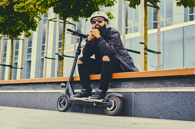 Stylischer bärtiger Mann mit Sonnenbrille, der sich auf einer Bank entspannt, nachdem er mit dem Elektroroller in der Innenstadt gefahren ist.