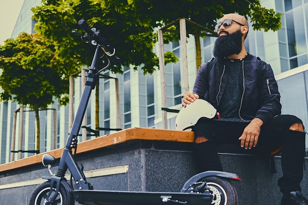 Stylischer bärtiger Mann mit Sonnenbrille, der sich auf einer Bank entspannt, nachdem er mit dem Elektroroller in der Innenstadt gefahren ist.