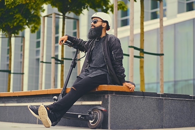 Stylischer bärtiger Mann mit Sonnenbrille, der sich auf einer Bank entspannt, nachdem er mit dem Elektroroller in der Innenstadt gefahren ist.