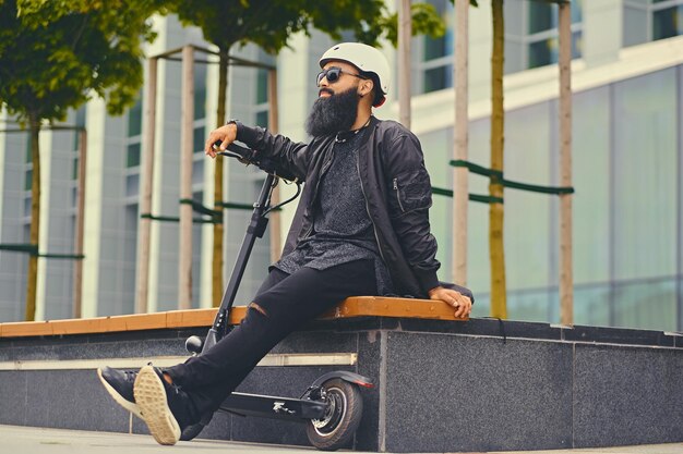 Stylischer bärtiger Mann mit Sonnenbrille, der sich auf einer Bank entspannt, nachdem er mit dem Elektroroller in der Innenstadt gefahren ist.