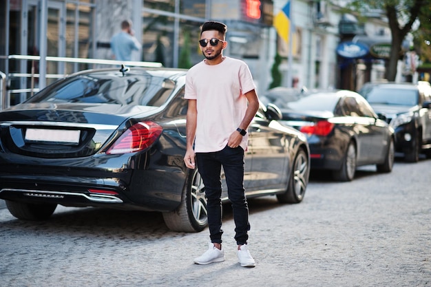 Stylischer indischer Bartmann mit Sonnenbrille und rosafarbenem T-Shirt gegen Luxusauto Indiens reiches Modell posierte im Freien auf den Straßen der Stadt