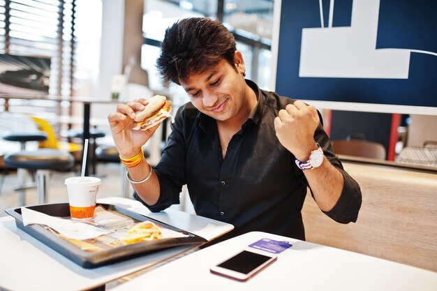 Stylischer indischer Mann, der im Fast-Food-Café sitzt und Hamburger isst, liest Morgennachrichten auf dem Handy. Er ist glücklich und zeigt, wie er etwas gewinnt