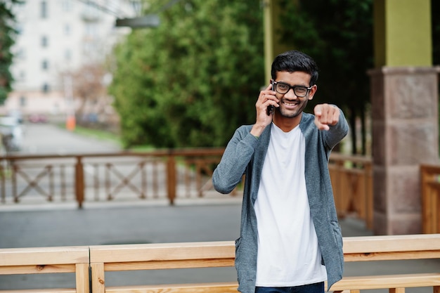 Stylischer indischer Mann mit Brille trägt lässige Posen im Freien und spricht am Telefon. Zeigen Sie den Finger zur Kamera