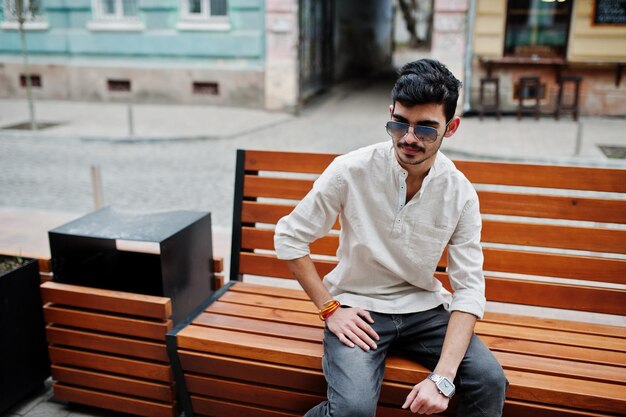 Stylischer indischer Modellmann in Freizeitkleidung und Sonnenbrille posierte im Freien auf der Straße von Indien und saß auf einer Bank