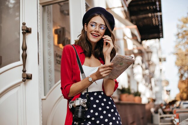 Stylischer junger Fotograf in schwarz-weißem und rotem Outfit mit brünetten, gewellten Haaren, Brille und Baskenmütze, die vor sonnigem Stadthintergrund stehen und wegschauen