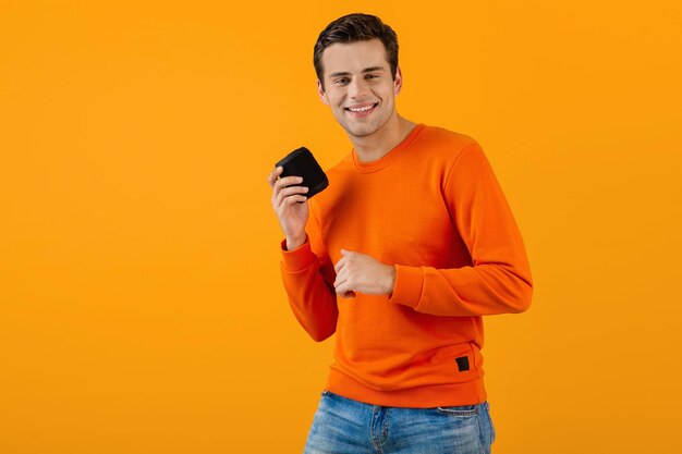 Stylischer lächelnder junger Mann im orangefarbenen Pullover mit drahtlosem Lautsprecher, der glücklich Musik hört und dabei fröhliche Stimmung im farbenfrohen Stil isoliert auf gelbem Hintergrund hat
