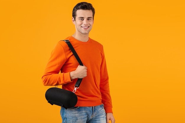 Stylischer lächelnder junger Mann im orangefarbenen Pullover mit drahtlosem Lautsprecher, der glücklich Musik hört und dabei fröhliche Stimmung im farbenfrohen Stil isoliert auf gelbem Hintergrund hat