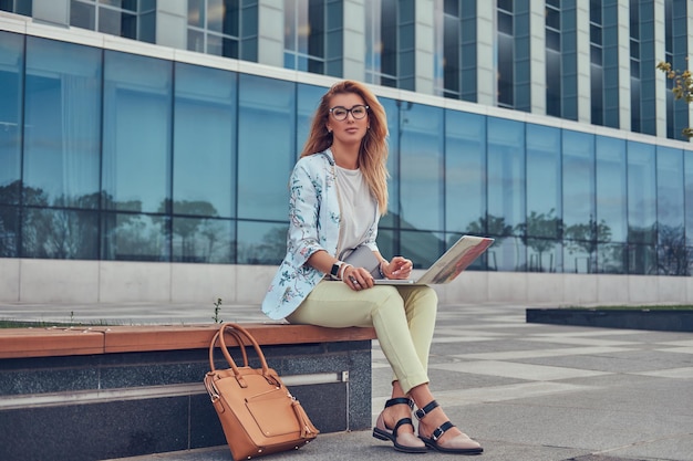 Stylischer Modeblogger entspannt sich im Freien, arbeitet am Laptop und sitzt auf einer Bank vor einem Wolkenkratzer.