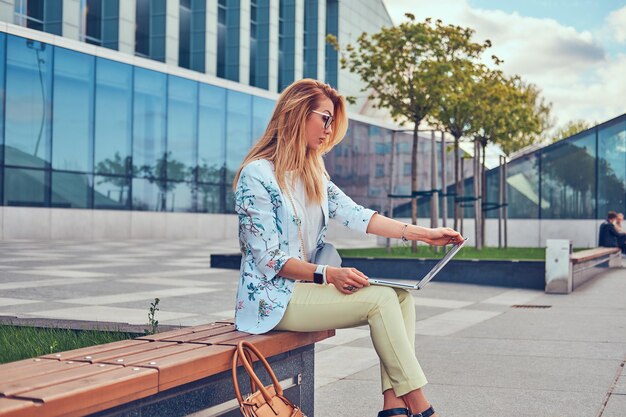 Stylischer Modeblogger entspannt sich im Freien, arbeitet am Laptop und sitzt auf einer Bank vor einem Wolkenkratzer.