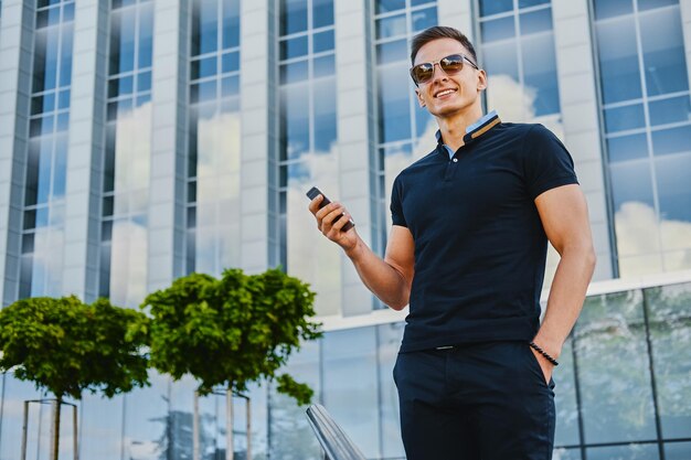 Stylischer muskulöser Mann mit Smartphone über modernem Gebäudehintergrund in der Innenstadt.