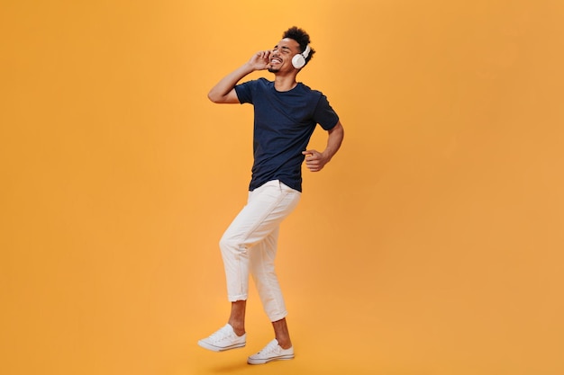 Stylischer Typ bewegt sich rhythmisch und genießt Musik über Kopfhörer Mann in weißer Hose und blauem Hemd tanzt auf orangefarbenem Hintergrund