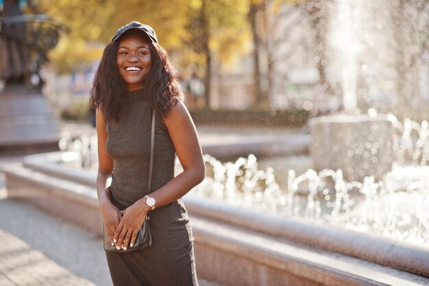 Stylisches afrikanisch-amerikanisches Mädchen in grauer Tunika-Crossbody-Tasche und Mütze posierte am sonnigen Herbsttag gegen Springbrunnen Afrika-Modellfrau