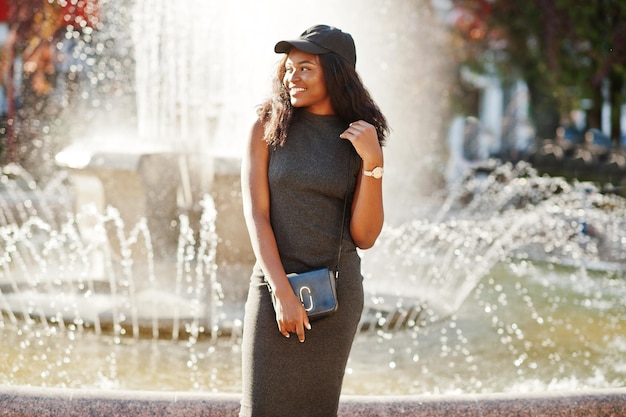 Stylisches afrikanisch-amerikanisches Mädchen in grauer Tunika-Crossbody-Tasche und Mütze posierte am sonnigen Herbsttag gegen Springbrunnen Afrika-Modellfrau