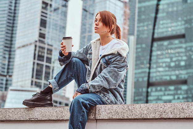 Stylisches rothaariges Hipster-Mädchen mit Tattoo auf ihrem Gesicht, das Jeansmantel trägt und Kaffee zum Mitnehmen hält, der am bewölkten Morgen vor Wolkenkratzern in der Stadt Moskau sitzt