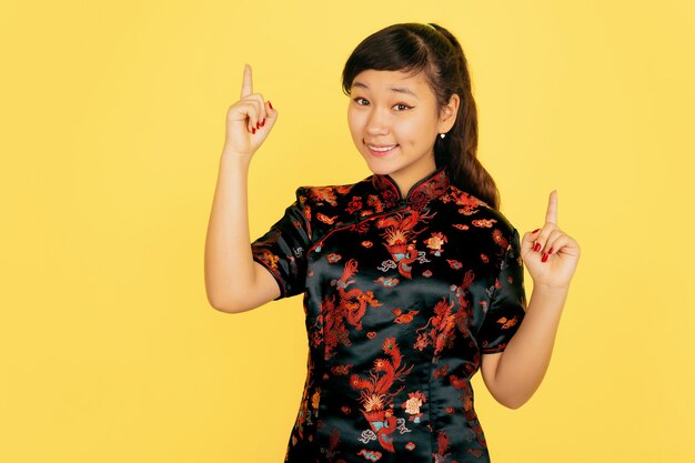 Süß lächelnd, nach oben zeigend. Frohes chinesisches neues Jahr 2020. Porträt des asiatischen jungen Mädchens auf gelbem Hintergrund. Weibliches Modell in traditioneller Kleidung sieht glücklich aus. Feier, menschliche Gefühle. Copyspace.