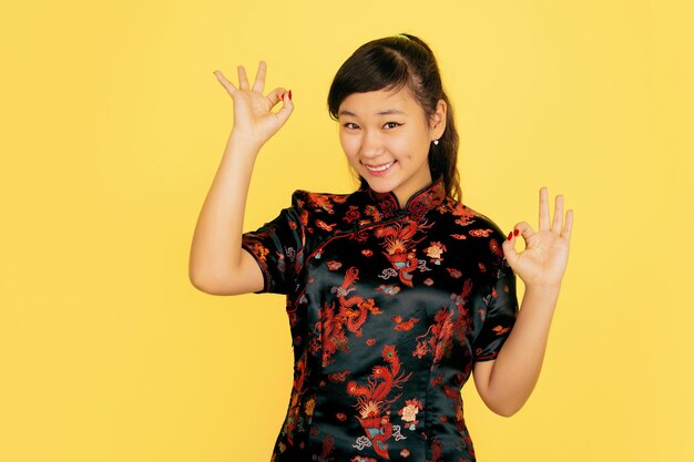 Süß lächelnd, nett zeigend. Frohes chinesisches Neujahr. Asiatisches junges Mädchenporträt auf gelbem Hintergrund. Weibliches Modell in traditioneller Kleidung sieht glücklich aus. Feier, menschliche Gefühle. Copyspace.