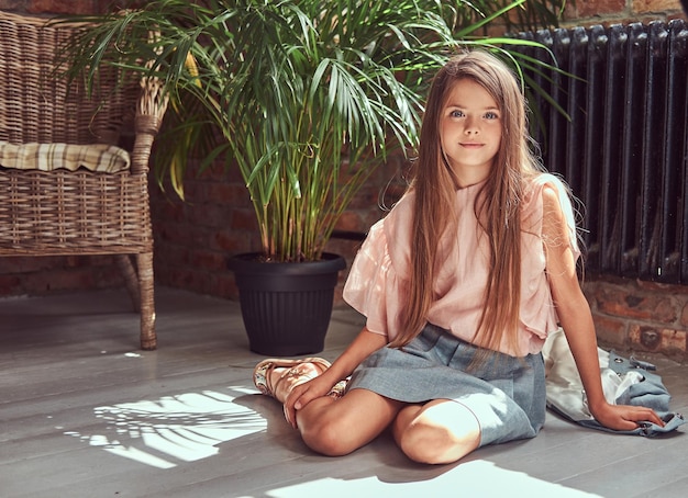 Süß lächelndes kleines Mädchen mit langen braunen Haaren, das ein stilvolles Kleid trägt und auf einem Holzboden in einem Raum mit Loft-Interieur sitzt.