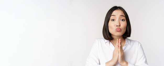 Süße asiatische Frau, die bittet, bitte um Gefallen zu bitten, braucht Hilfe, wenn sie mit schüchternem Gesicht vor weißem Hintergrund steht