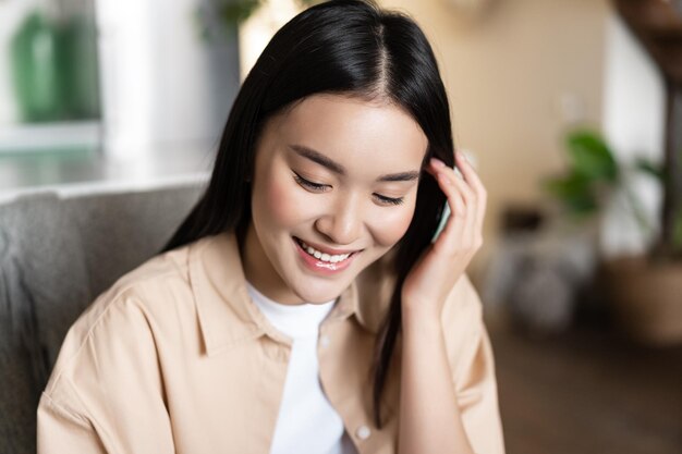 Süße asiatische Studentin lächelnd nach unten schauen und Haare hinter dem Ohr stecken zu Hause drinnen sitzen in ...