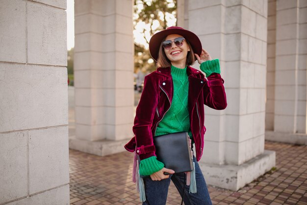 Süße Frau im trendigen Outfit im Herbststil, die auf der Straße mit lila Samtjacke, Sonnenbrille und Hut spazieren geht