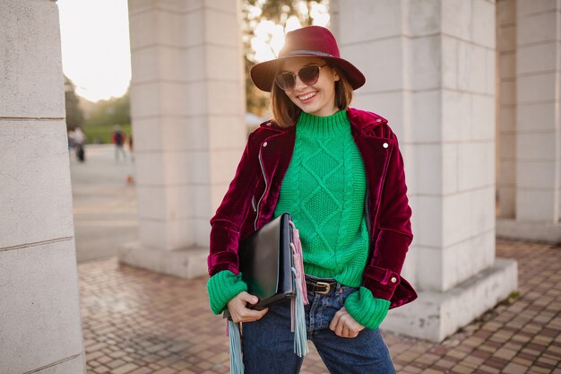 Süße Frau im trendigen Outfit im Herbststil, die auf der Straße mit lila Samtjacke, Sonnenbrille und Hut spazieren geht