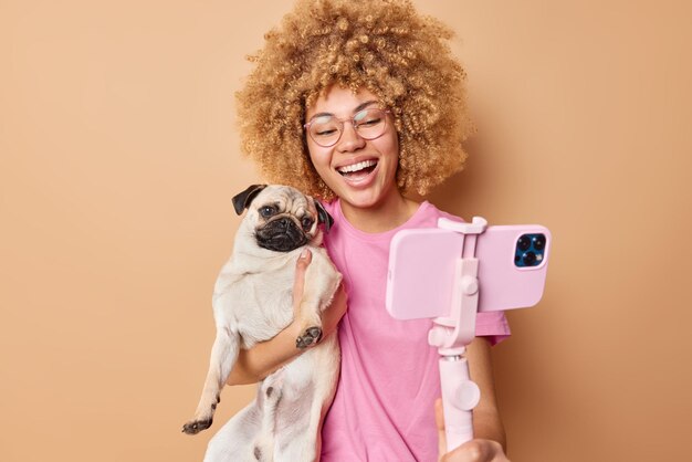 Süße, fröhliche Tierhalterin posiert mit Pug-Hund für Selfie, verbringen Freizeit zusammen und machen ein unvergessliches Foto, während sie über braunem Hintergrund spazieren gehen Fröhliche junge Frau mit Haustier