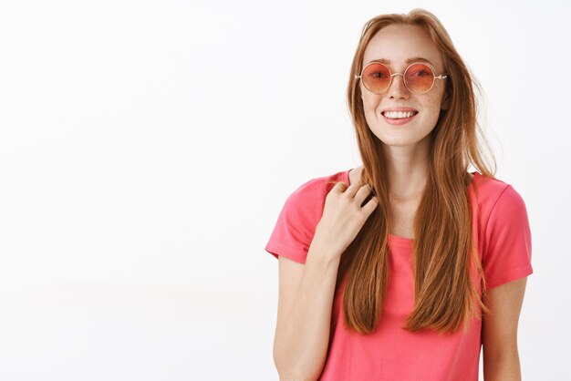 süße fröhliche und emotionale junge rothaarige Frau in rosa Sonnenbrille und T-Shirt, die Haarsträhne berührt und lächelt, fühlt sich unbehaglich in unbekannter Gesellschaft, die versucht, Gespräch freundlich zu beginnen