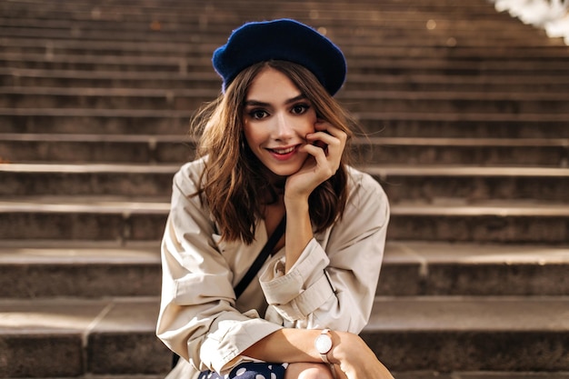 Süße junge Dame in stylischem beige Trenchcoat mit blauer Baskenmütze und roten Lippen lächelt und posiert auf alten Treppen im Freien und sieht direkt aus Sonniger Herbst