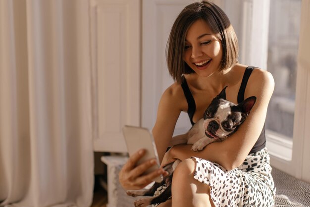 Süße junge kaukasische Brünette umarmt Hund mit Vergnügen und macht Selfie am Telefon Liebe für Tiere Haustierkonzept