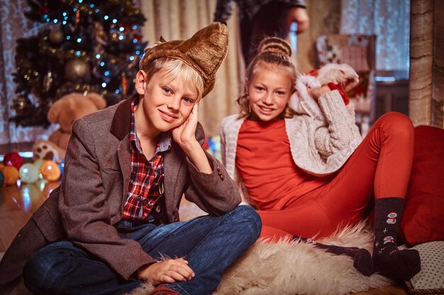 Süße kleine Teenager, glücklicher Bruder und Schwester, die zu Hause auf einem Pelzteppich neben einem Weihnachtsbaum sitzen.