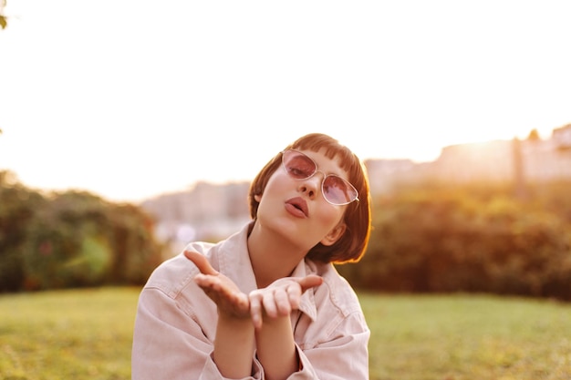 Süße kurzhaarige Frau bläst Kuss, ich parke Attraktives charmantes Mädchen mit Sonnenbrille und rosa Jeansjacke posiert draußen