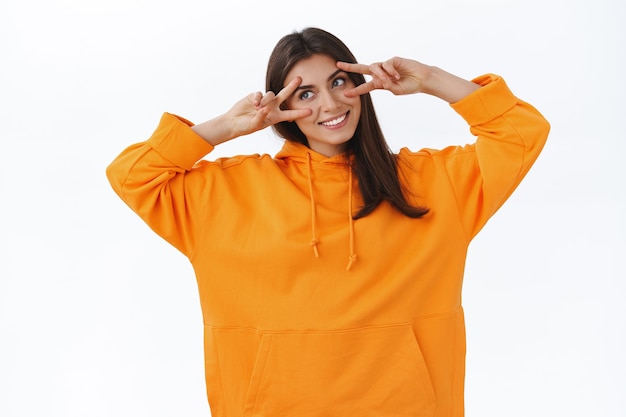 Süße zarte weibliche Kaukasierin in orangefarbenem Hoodie, zeigt Friedenszeichen über den Augen, neigt den Kopf und schaut glücklich nach links, lächelt breit und drückt positive Stimmung, Jubel und Freude aus, weiße Wand