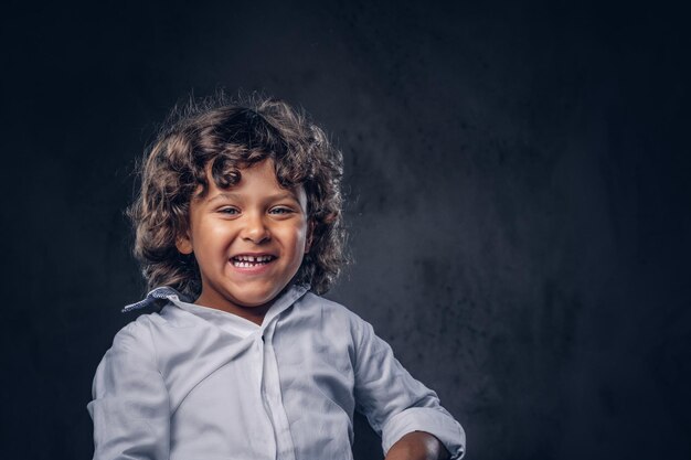 Süßer lächelnder Schuljunge mit braunem, lockigem Haar, gekleidet in ein weißes Hemd, posiert in einem Studio. Getrennt auf einem dunklen strukturierten Hintergrund.