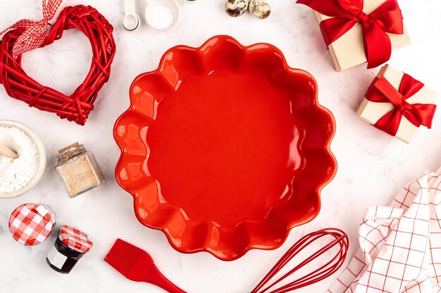 Süßes Backkonzept zum Valentinstag, Kochhintergrund mit Backen - Schneebesen zum Schlagen, rote Keramikkuchenform, Zucker, Mehl, Backpulver. Geschenkboxen mit Schleifen. Weißer Hintergrund, Ansicht von oben