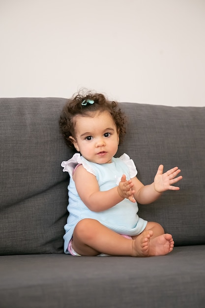 Süßes dunkles lockiges Baby in hellblauem Stoff, der auf grauer Couch zu Hause sitzt und Hände klatscht. Kinder zu Hause und Kindheitskonzept
