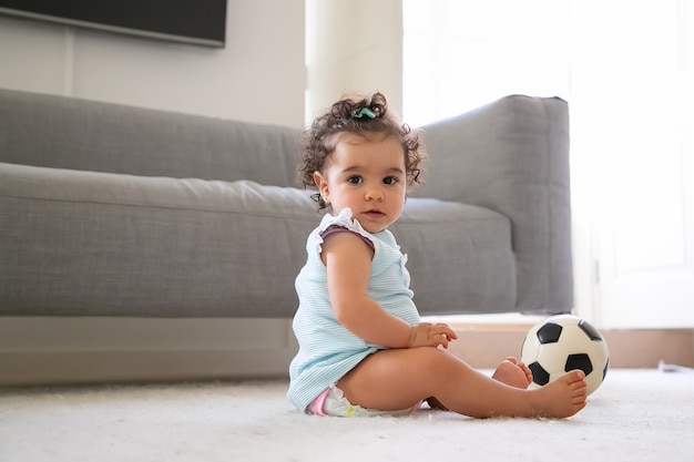 Süßes ernstes schwarzhaariges Baby in hellblauen Kleidern, die auf Boden mit Fußball sitzen, a. Seitenansicht. Kinder zu Hause und Kindheitskonzept